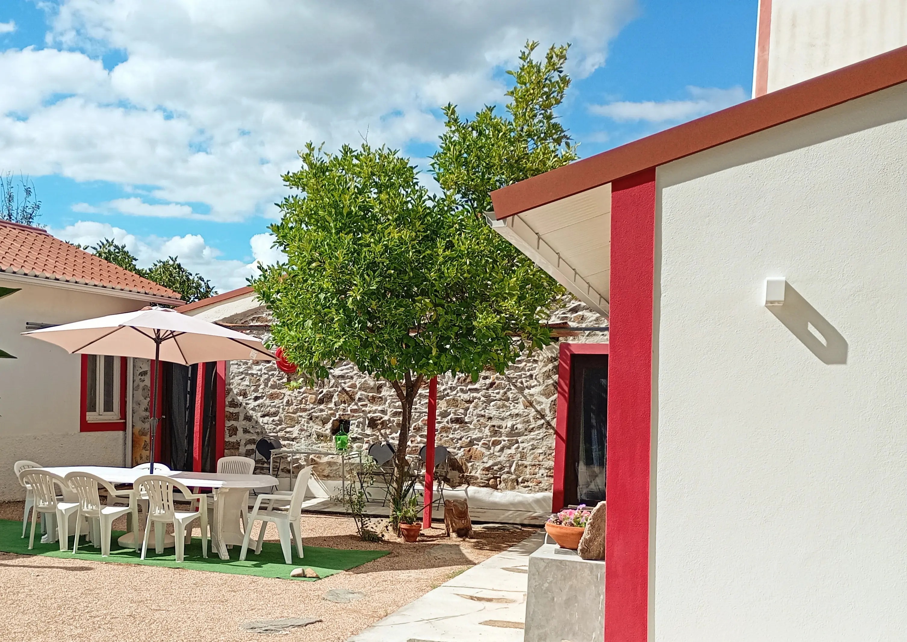 Pátio exterior com mesa e chapéu de sol, laranjeira ao centro e os remates vermelhos da casa