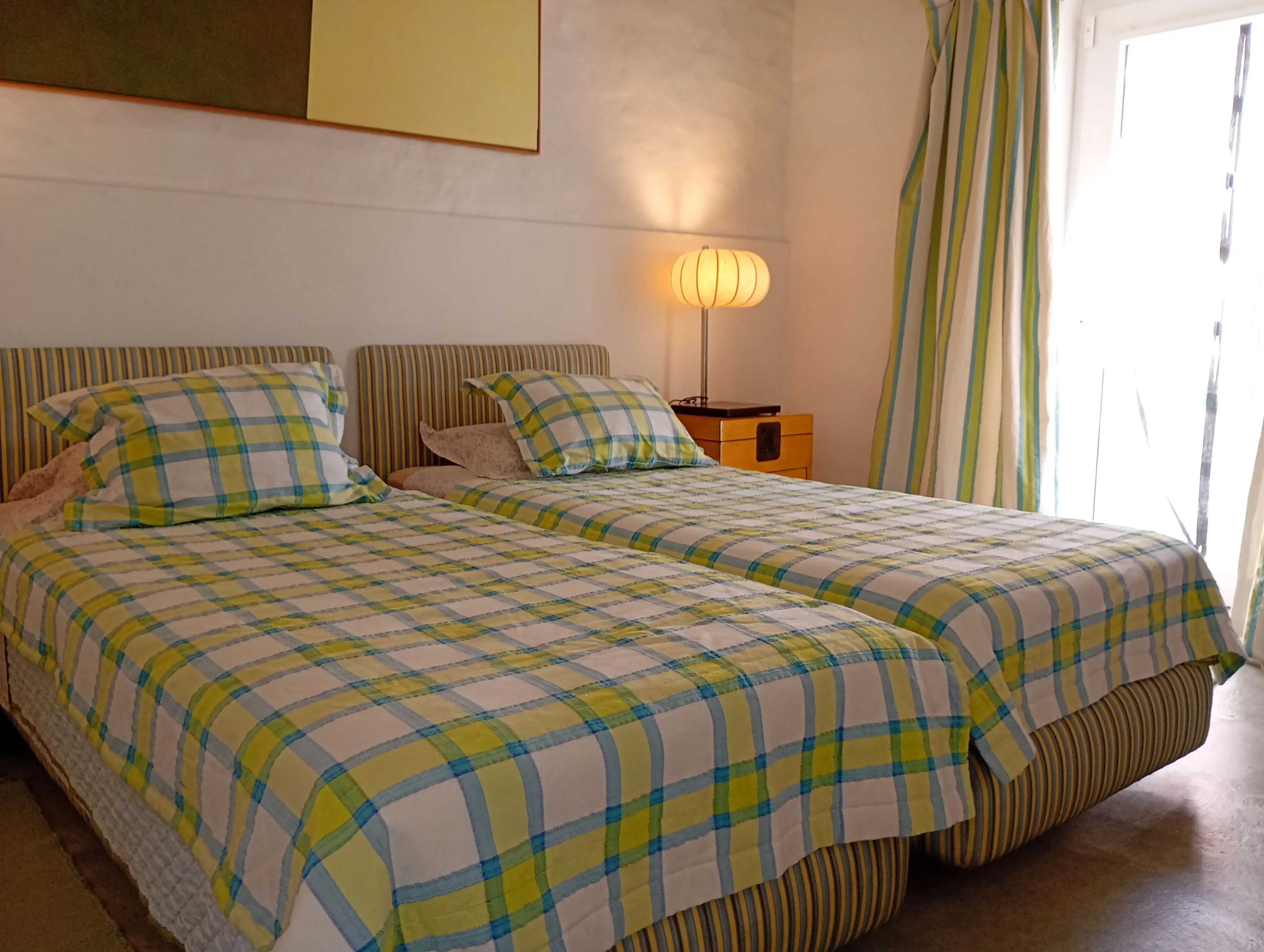 Quarto da Casa Principal com duas camas individuais, colchas aos quadrados verdes e azuis e porta para o exterior